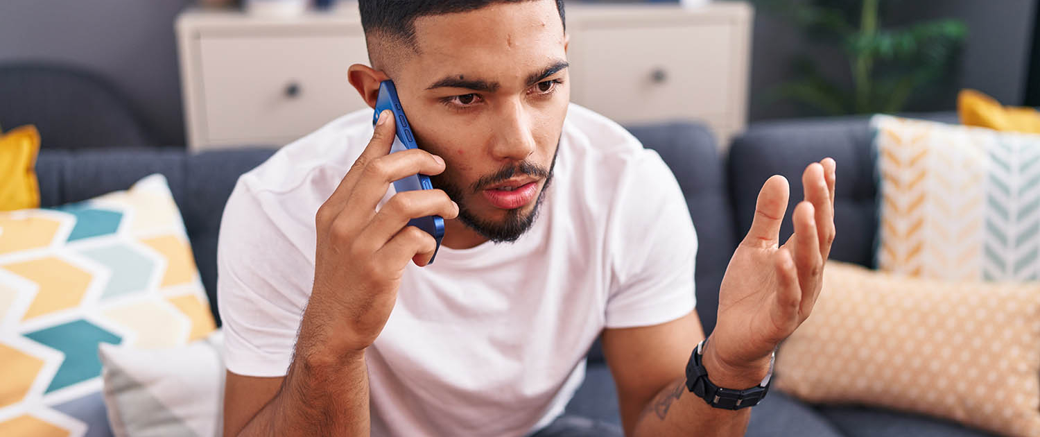 Young latin man talking on the smartphone sitting on sofa at home